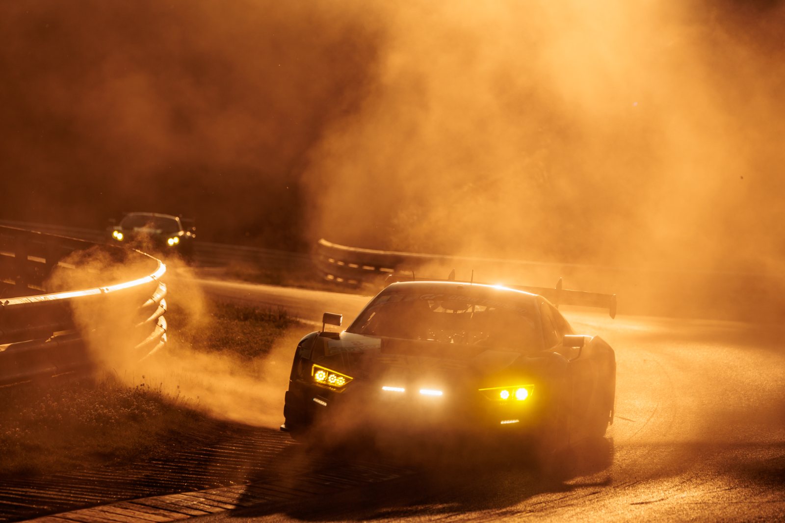 GT3-Rennwagen bei Nacht im Staub der Nordschleife, Scheinwerfer im Gegenlicht, Verfolger in der Kurve dahinter