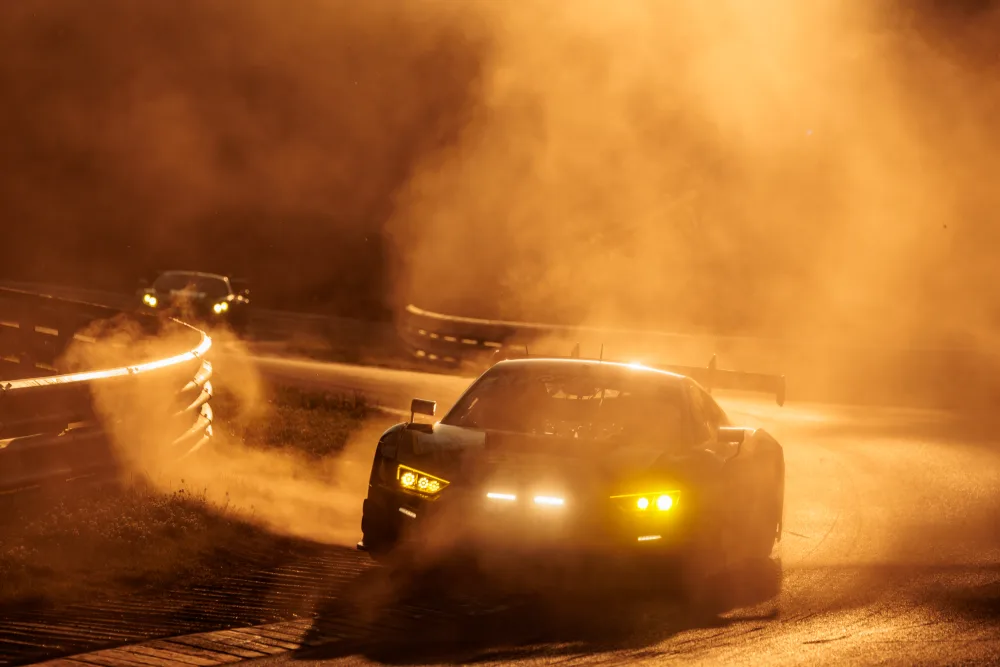 GT3-Rennwagen bei Nacht in aufgewirbeltem Staub, Scheinwerfer im Gegenlicht, dahinter ein zweites Fahrzeug