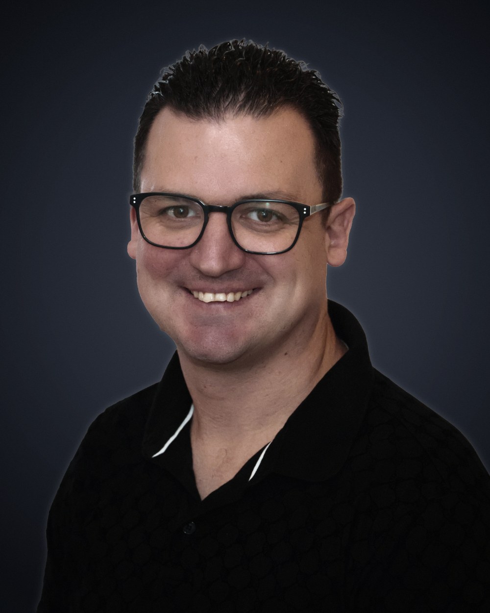 Patrick Liepertz, lächelnd, mit schwarzer Brille und schwarzem Polohemd vor dunkelgrauem Studiohintergrund