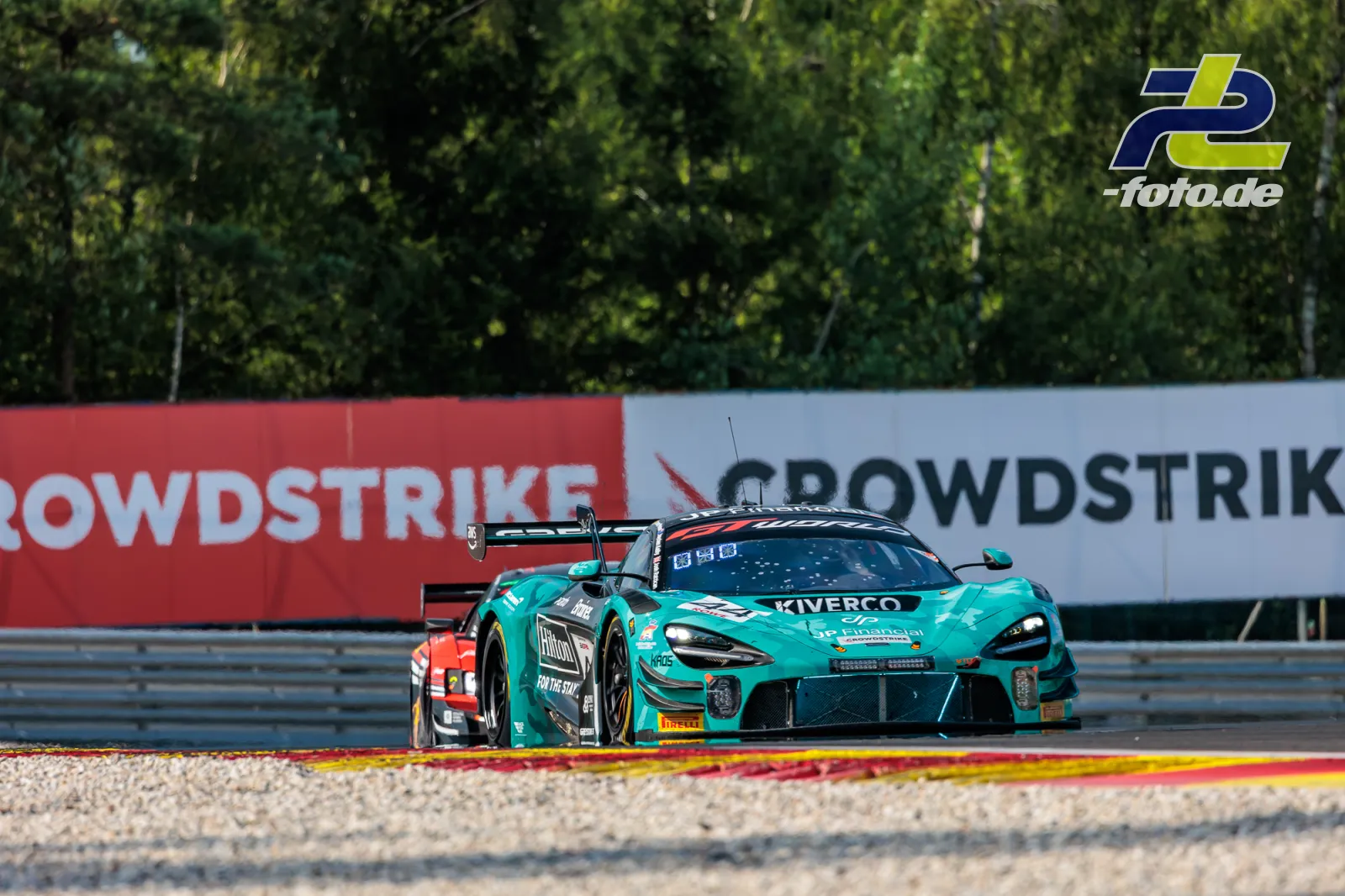 Türkisfarbener McLaren 720S GT3 EVO auf der Strecke beim 24-Stunden-Rennen von Spa-Francorchamps, im Hintergrund Streckenwerbung und Bäume.
