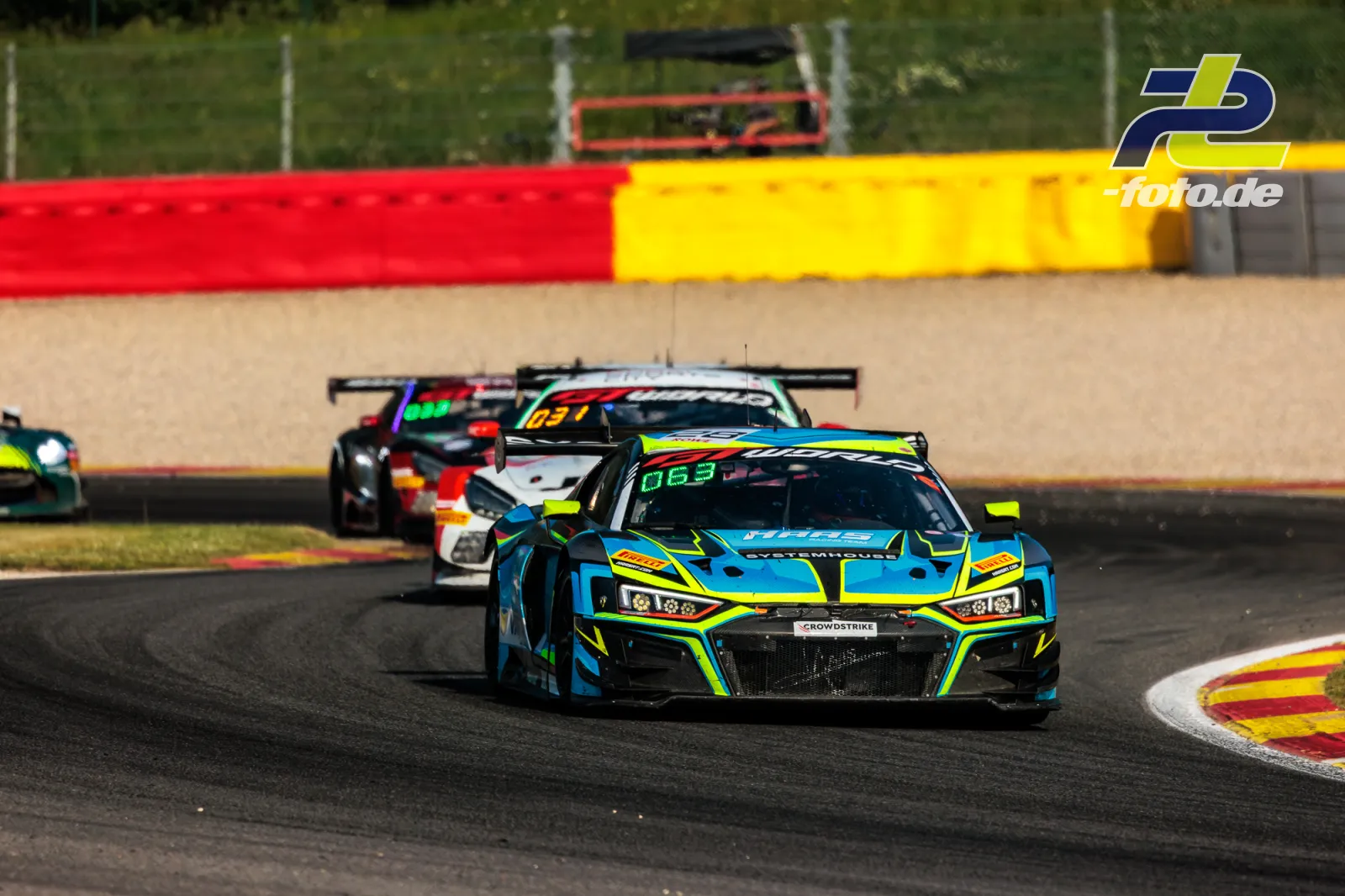 Audi R8 LMS GT3 EVO II #28 von Haas RT in blau-gelb-grüner Lackierung führt eine Gruppe GT3-Fahrzeuge durch eine Kurve in Spa-Francorchamps.