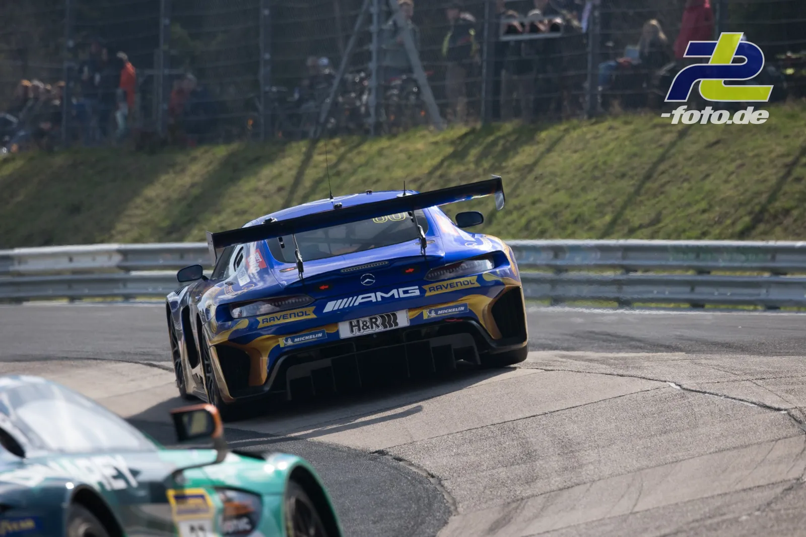 Mercedes-AMG GT3 in blau-gold auf der Nordschleife, verfolgt von einem Audi R8 LMS, Zuschauer am Streckenrand