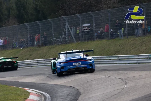 Blauer Porsche 911 GT3 Cup im Rennen auf dem Nürburgring, gefolgt von grünem Konkurrenten, Zuschauer im Hintergrund.