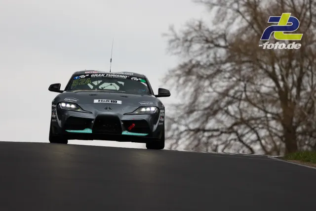 Toyota GR Supra mit Startnummer 524 über der Kuppe am Nürburgring, vor bewölktem Himmel und kahlen Bäumen