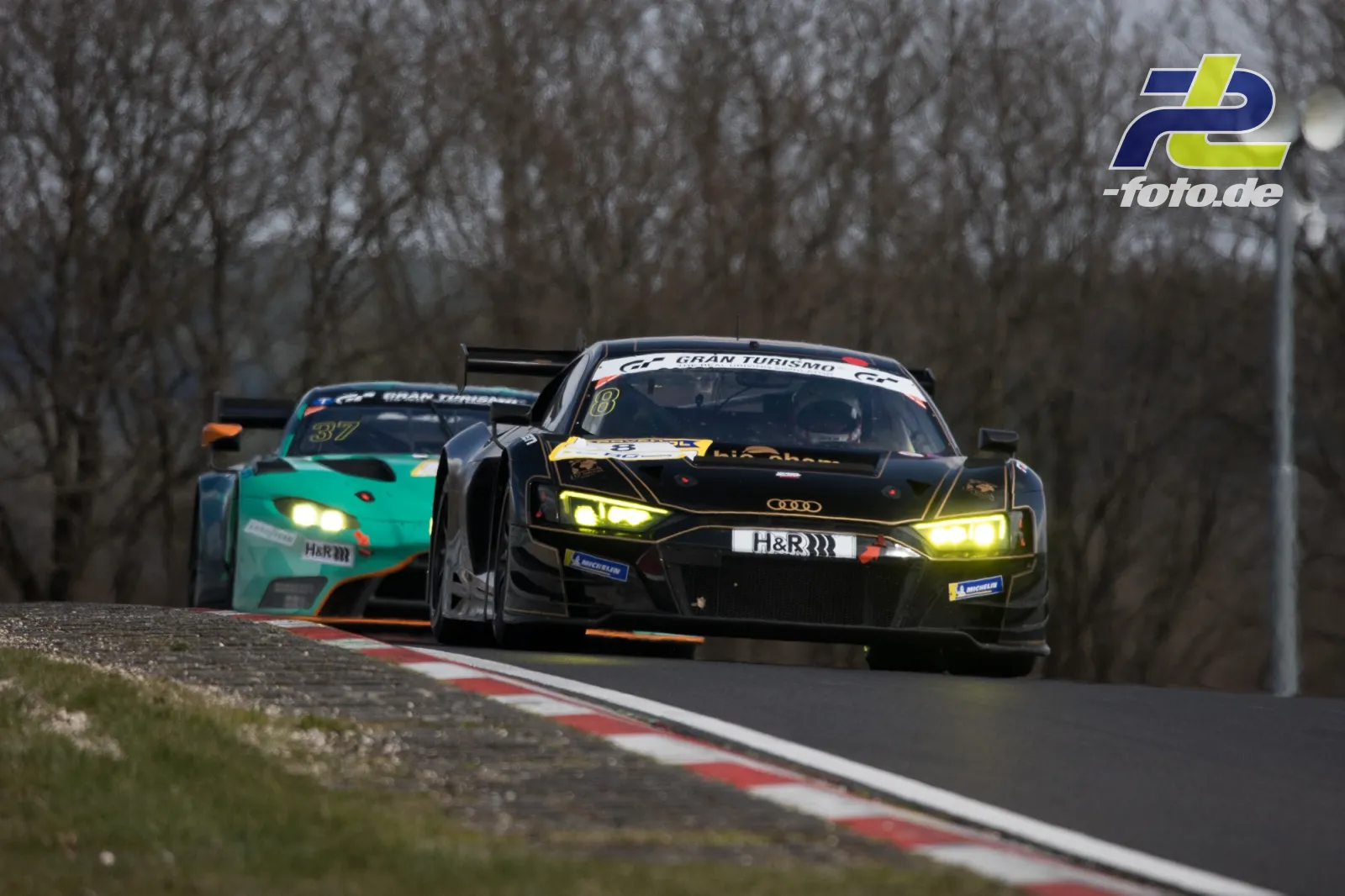 Zwei GT-Rennwagen mit eingeschaltetem Licht in Kurve auf der Nordschleife bei bedecktem Wetter, vorne Audi LMS GT3 mit H&R-Schriftzug.