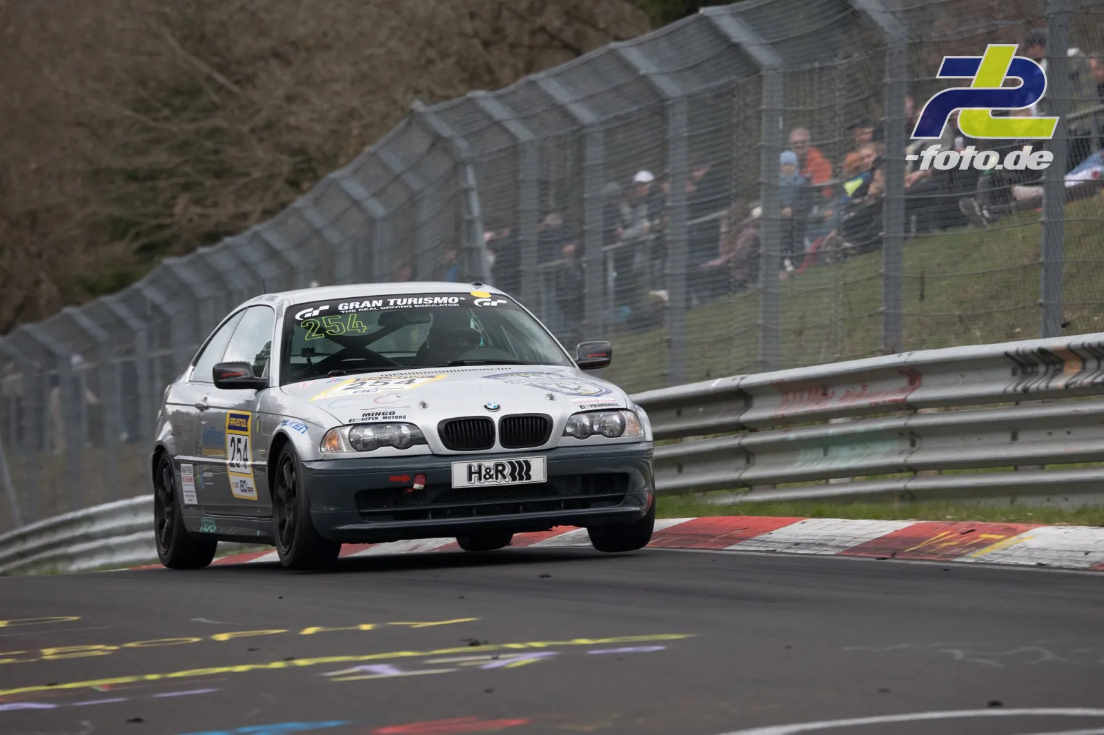 BMW E46 Nummer 254 auf der Nürburgring-Nordschleife, im Hintergrund Fangzaun und Zuschauer beim NLS-Rennen.