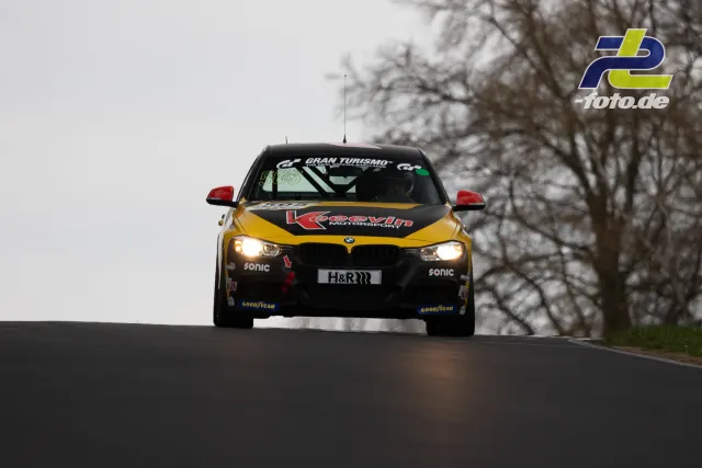 BMW-Rennwagen mit gelb-schwarzer Lackierung frontal auf der Nordschleife, Licht an, Bäume im Hintergrund