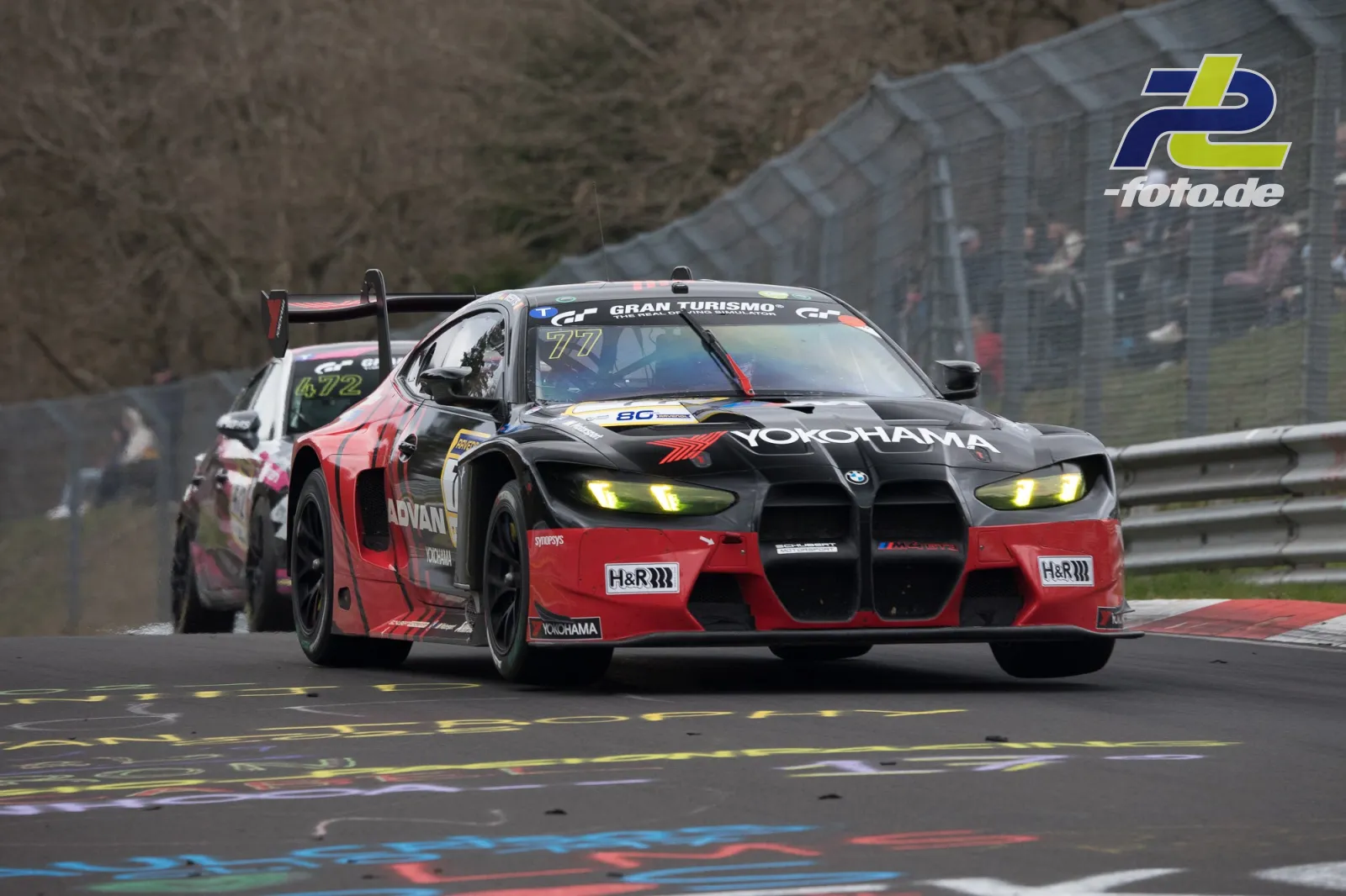 BMW M4 GT3 mit Startnummer 77 in rot-schwarzer Lackierung auf nasser Nordschleife beim NLS-Rennen, verfolgt von einem weiteren GT-Fahrzeug.