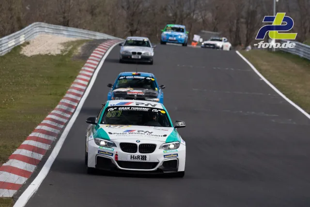 Rennszene auf der Nordschleife mit mehreren Fahrzeugen, angeführt von einem weißen BMW mit türkisfarbenen Akzenten