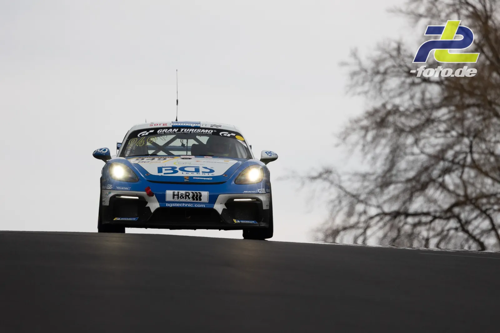 Porsche Cayman GT4 springt über Kuppe auf der Nordschleife, Frontansicht mit eingeschalteten Scheinwerfern bei trübem Wetter.