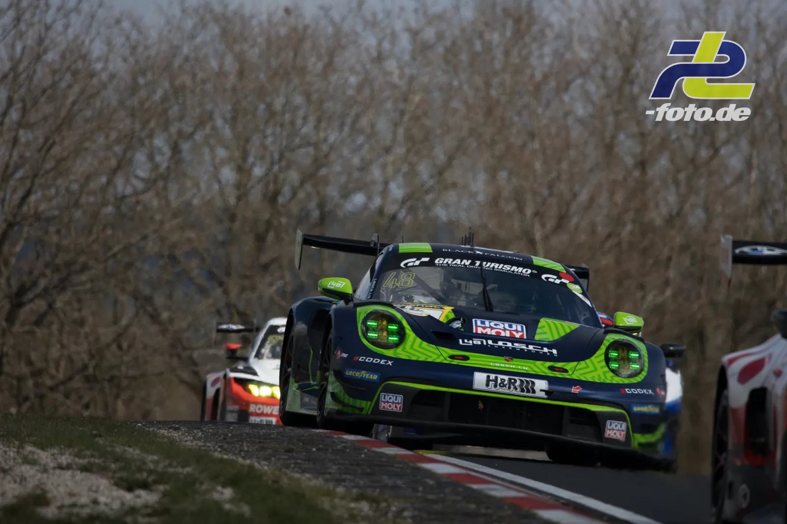 Porsche 911 GT3 R in schwarz-grüner Lackierung führt ein Feld durch eine Kurve auf dem Nürburgring. Weitere GT3-Fahrzeuge folgen.