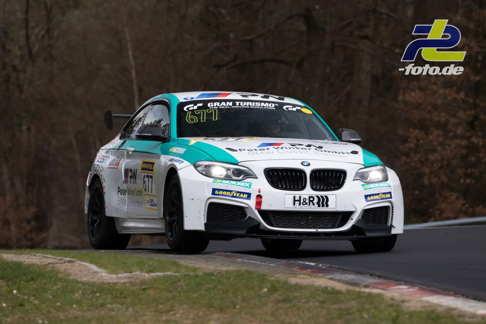BMW M240i Nummer 677 in weiß-türkis beim NLS-Rennen auf der Nürburgring-Nordschleife in Kurvenfahrt mit eingeschalteten Scheinwerfern.