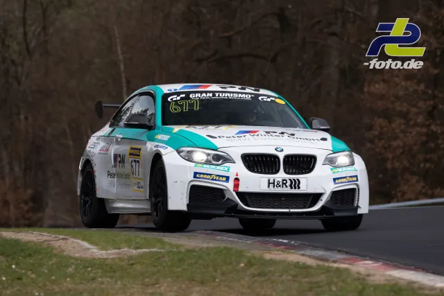 BMW M240i Nummer 677 in weiß-türkis beim NLS-Rennen auf der Nürburgring-Nordschleife in Kurvenfahrt mit eingeschalteten Scheinwerfern.