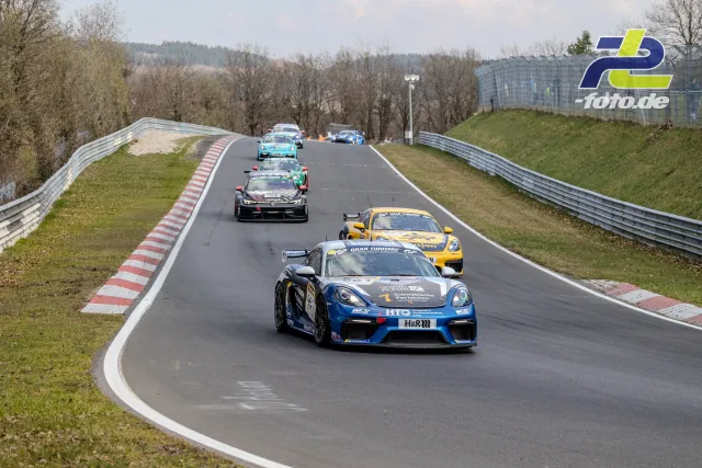 Fahrzeugfeld auf der Nordschleife während des Rennens, angeführt von einem blau lackierten Porsche Cayman.