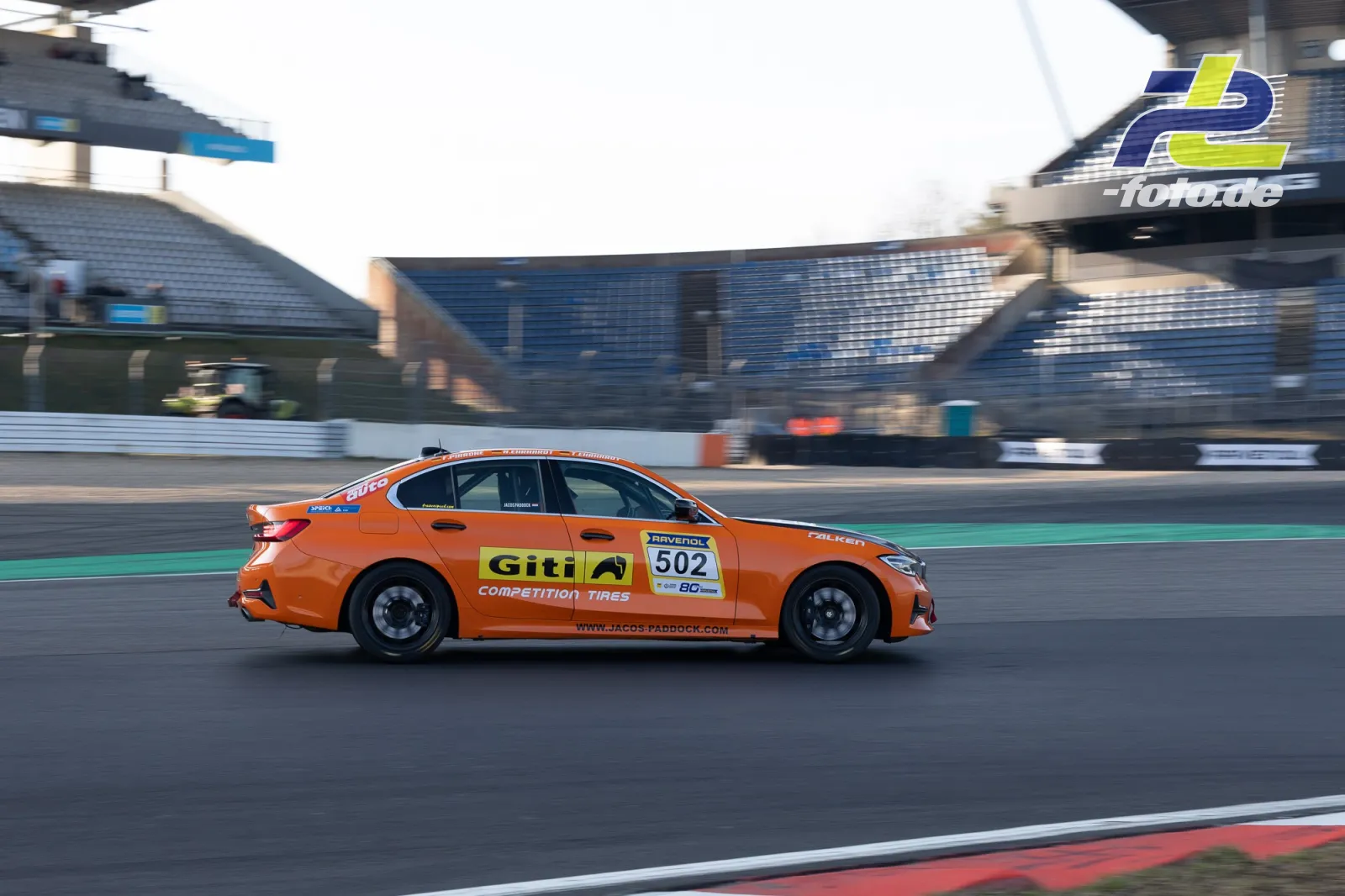Oranger BMW mit Startnummer 502 im Zeittraining auf dem Nürburgring, Mitzieher mit unscharfem Hintergrund und Tribüne.
