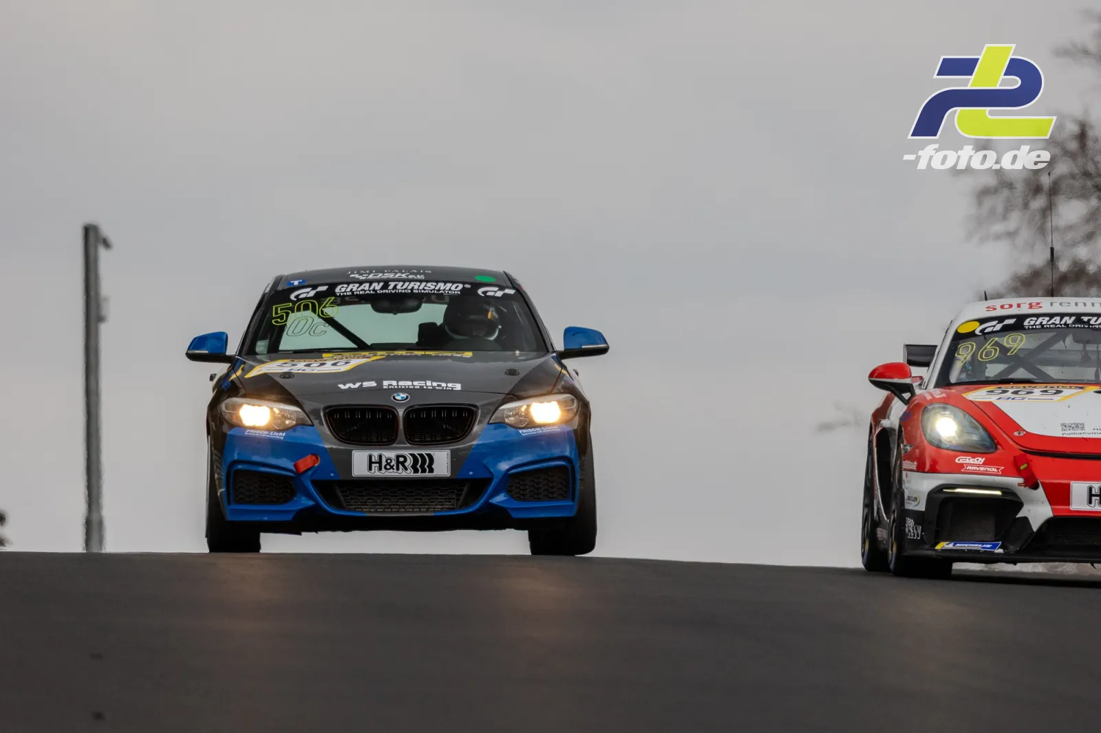 BMW und Porsche im Rennen auf der Nürburgring-Nordschleife bei trübem Wetter, Fahrzeuge mit eingeschaltetem Licht auf der Kuppe.
