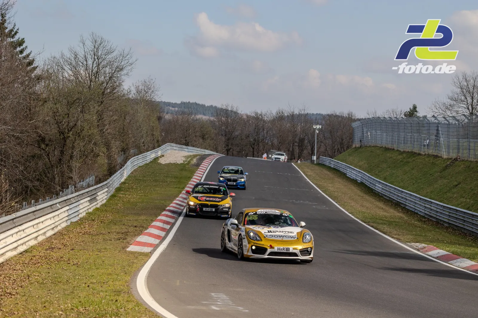 Vier Rennwagen auf der Nordschleife, angeführt von einem weiß-goldenen Porsche, der durch eine Kuppe fährt.