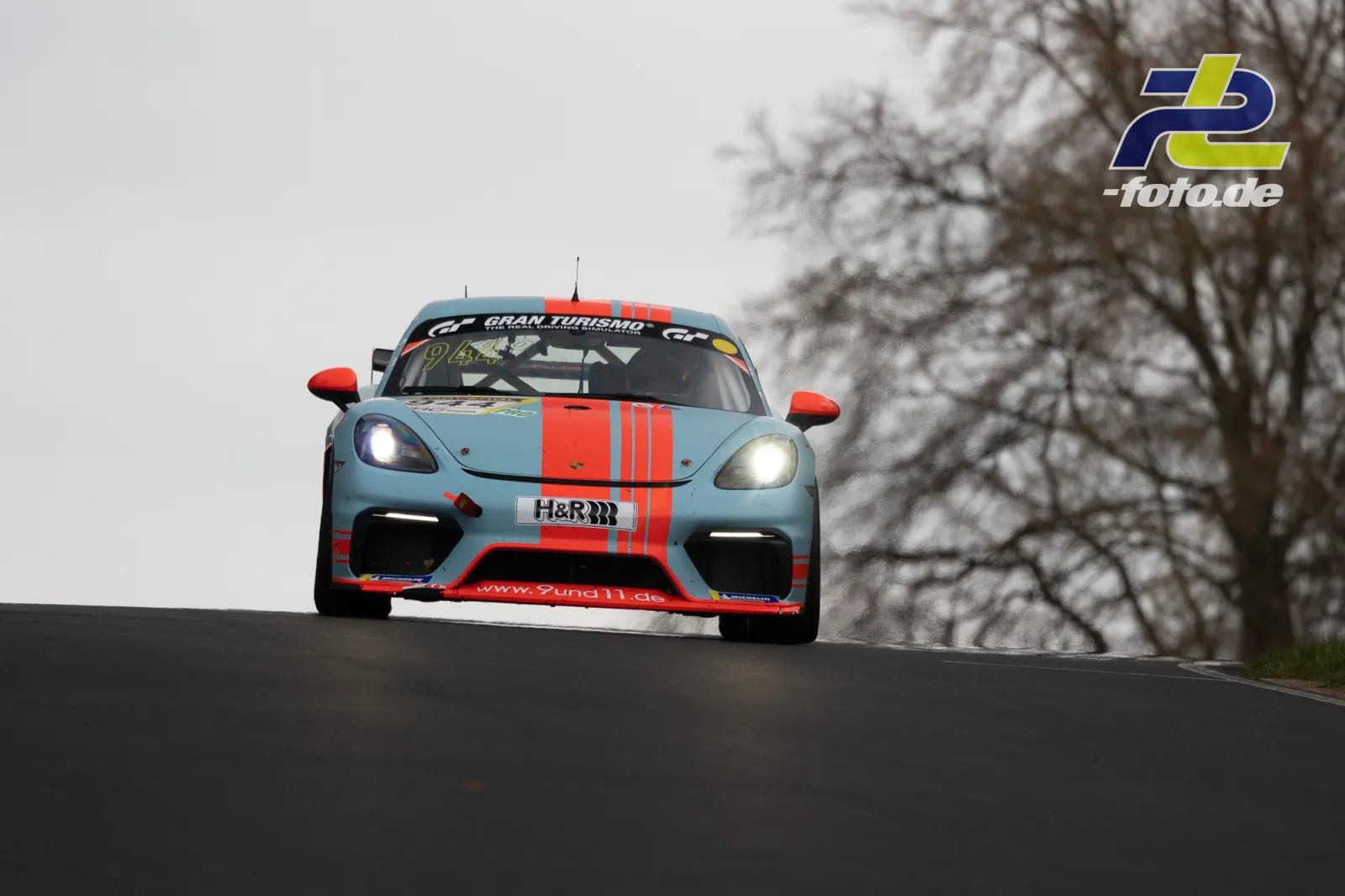 Porsche 718 Cayman GT4 Clubsport in hellblau-orange springt über eine Kuppe beim NLS-Lauf am Nürburgring.