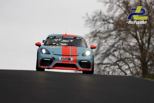 Porsche 718 Cayman GT4 Clubsport in hellblau-orange springt über eine Kuppe beim NLS-Lauf am Nürburgring.