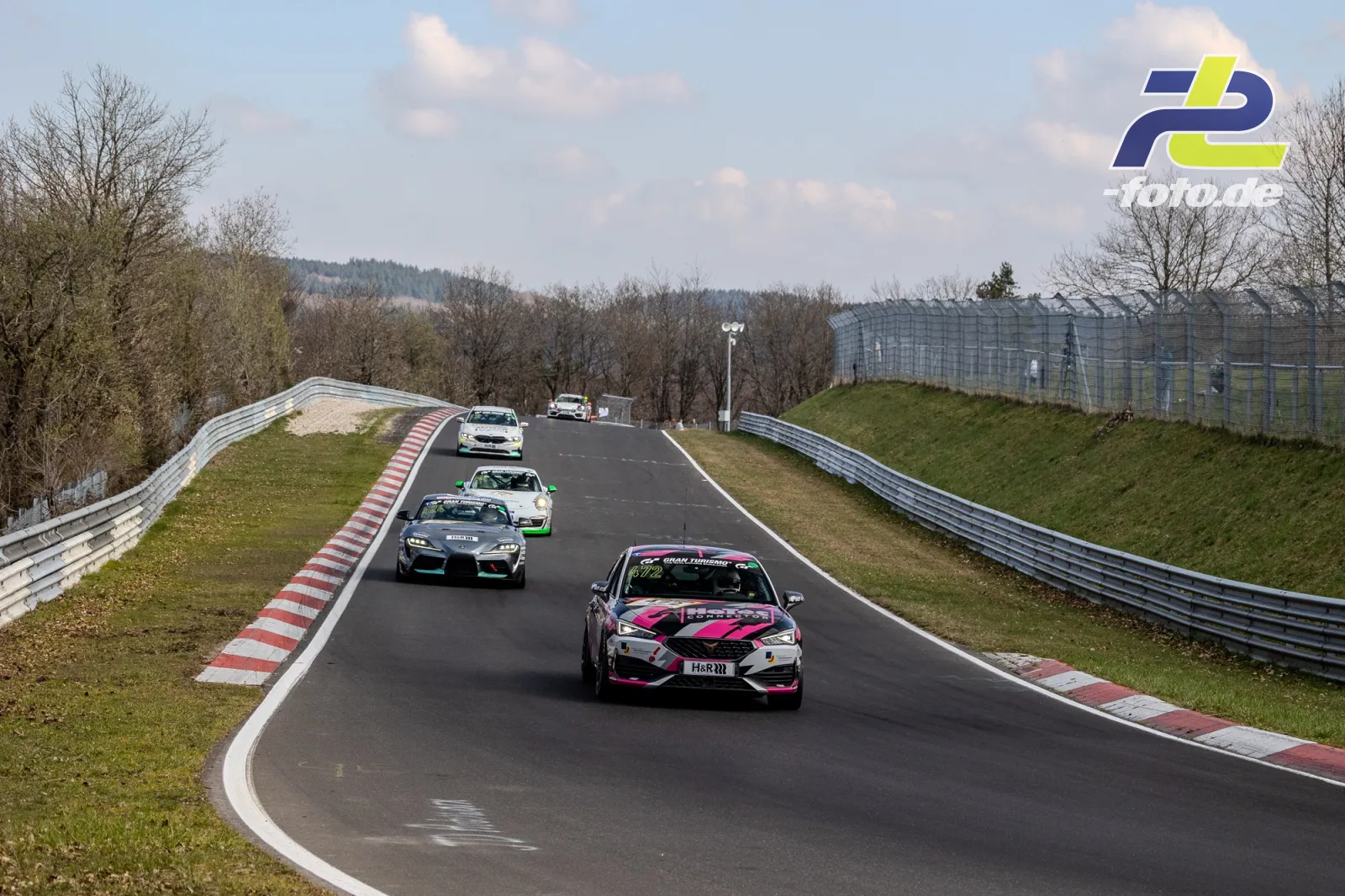 Mehrere Fahrzeuge im Rennen auf der Nordschleife, angeführt von einem pink-weißen Tourenwagen, gefolgt von weiteren Fahrzeugen auf einer Kuppe.
