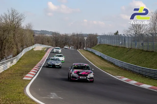 Mehrere Fahrzeuge im Rennen auf der Nordschleife, angeführt von einem pink-weißen Tourenwagen, gefolgt von weiteren Fahrzeugen auf einer Kuppe.