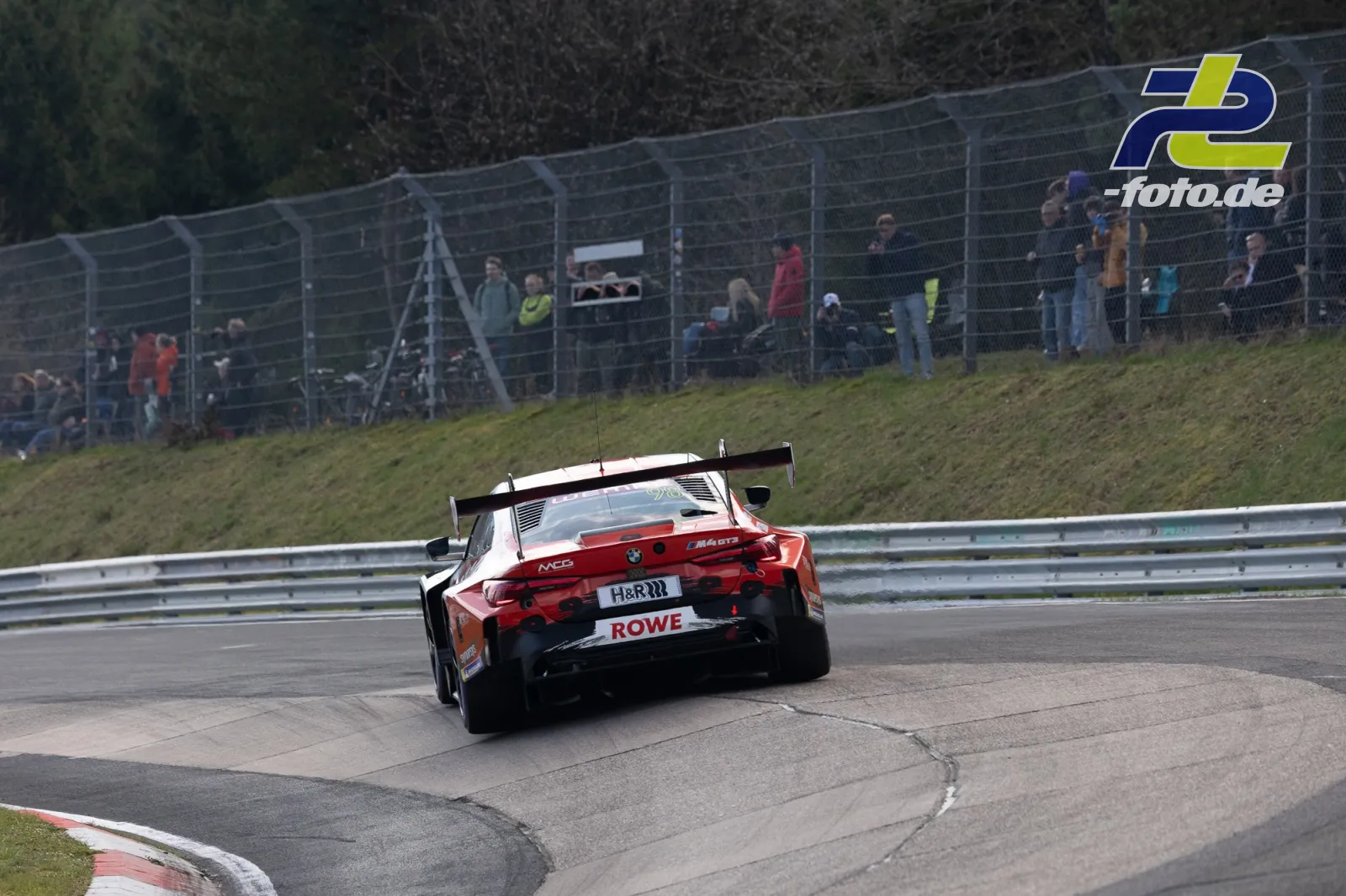 BMW M4 GT3 in rot-weißer Lackierung auf der Nordschleife, Zuschauer am Streckenzaun