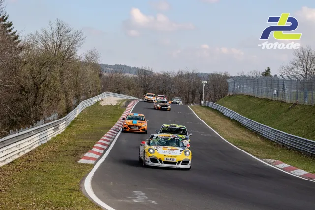 Rennfeld auf der Nürburgring-Nordschleife mit gelbem Porsche 911 an der Spitze, gefolgt von Tourenwagen in einer Rechtskurve.