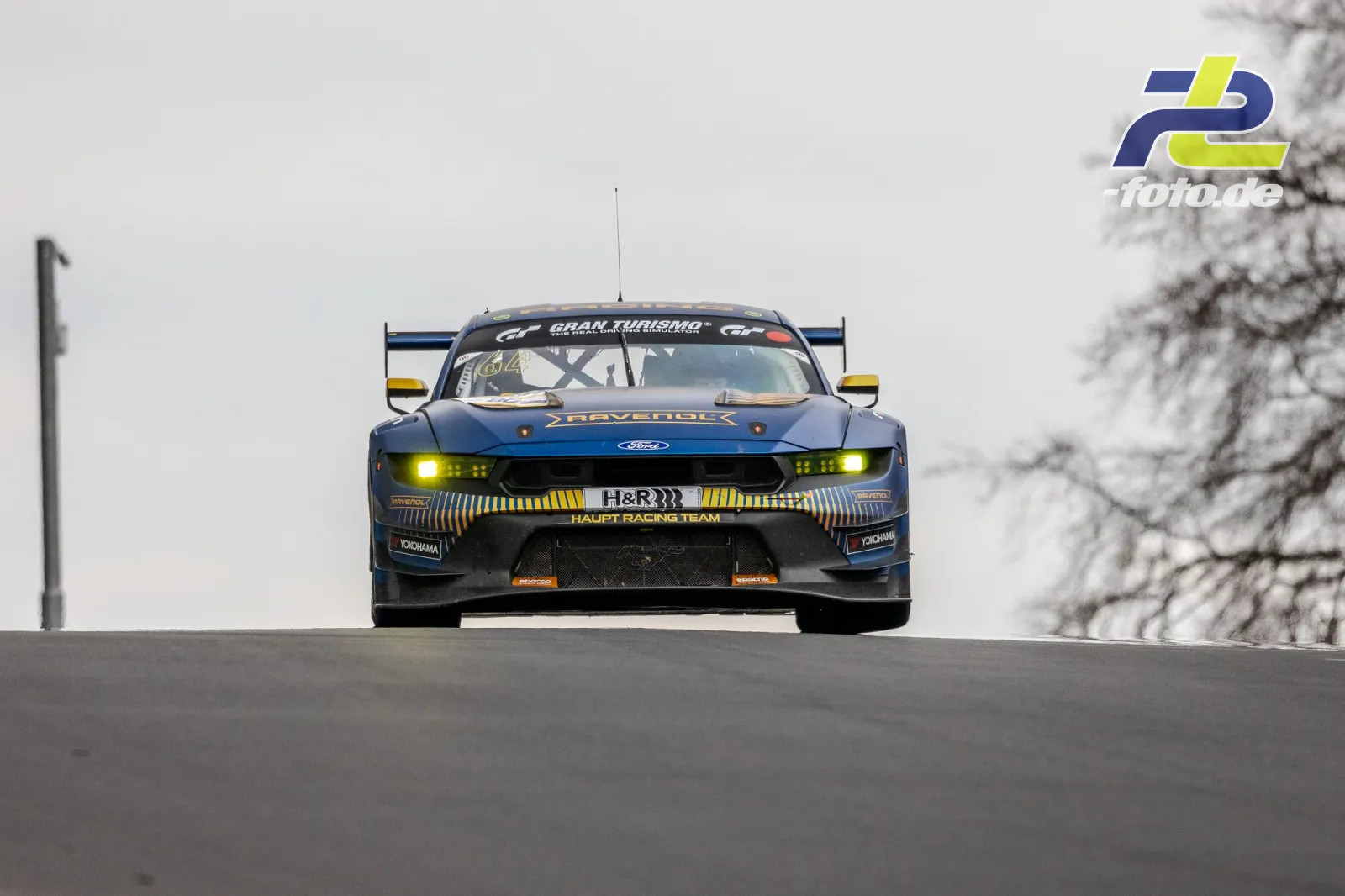 Ford Mustang GT4 von Haupt Racing Team über Kuppe auf der Nordschleife mit eingeschalteten Scheinwerfern bei bedecktem Himmel.