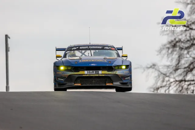 Ford Mustang GT4 von Haupt Racing Team über Kuppe auf der Nordschleife mit eingeschalteten Scheinwerfern bei bedecktem Himmel.