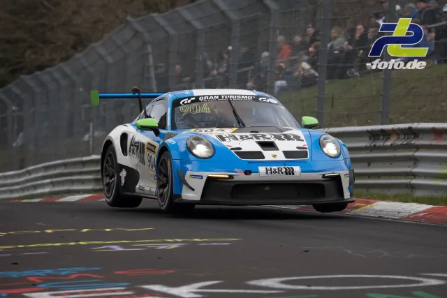 Porsche 911 GT3 R Nr. 904 in hellblau-weißer Lackierung auf der Nordschleife beim NLS-Rennen, Zuschauer an der Tribüne.