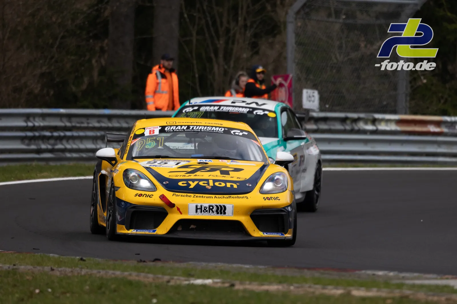 Zwei Rennwagen auf der Nordschleife, vorn ein gelber Porsche mit Startnummer 951, Streckenposten im Hintergrund.