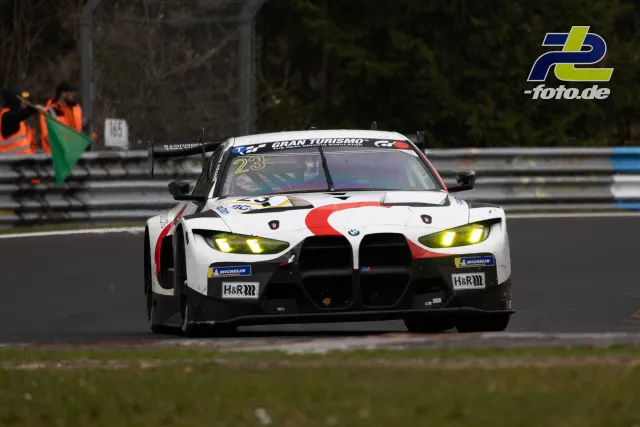 BMW M4 GT3 mit Startnummer 46 frontal auf der Nordschleife, eingeschaltete Scheinwerfer, Streckenposten mit grüner Flagge im Hintergrund.
