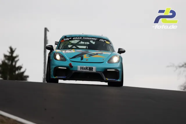 Türkiser Porsche Cayman GT4 mit Gran Turismo-Schriftzug über eine Kuppe springend, Nürburgring Nordschleife NLS-Rennen.