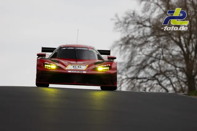 Roter Rennwagen mit gelben Scheinwerfern fährt über eine Kuppe auf der Nordschleife bei grauem Wetter.