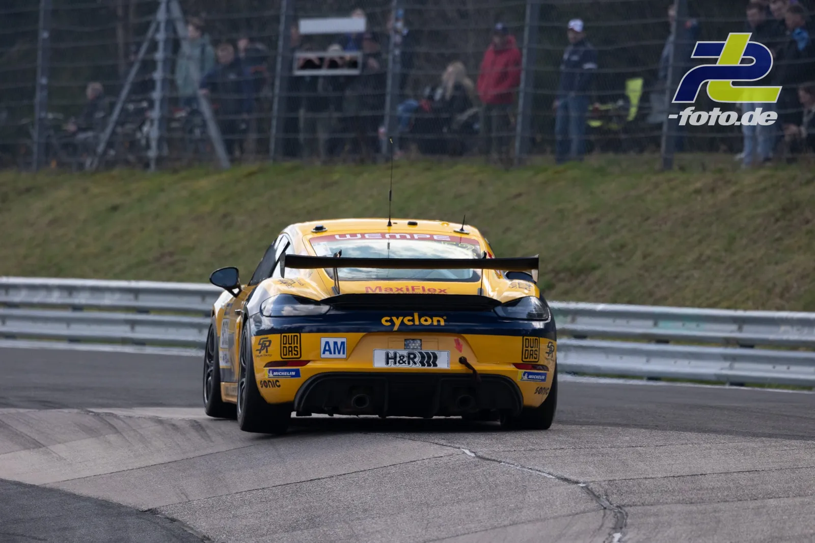 Porsche 718 Cayman GT4 in Gelb-Schwarz aus Heckperspektive auf dem Nürburgring bei NLS-Rennen, Zuschauer im Hintergrund.