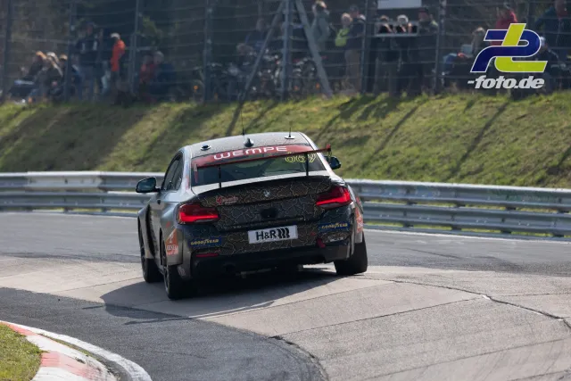 BMW Renntourenwagen mit Startnummer 962 in Kurve auf dem Nürburgring, Zuschauer im Hintergrund hinter Fangzaun.