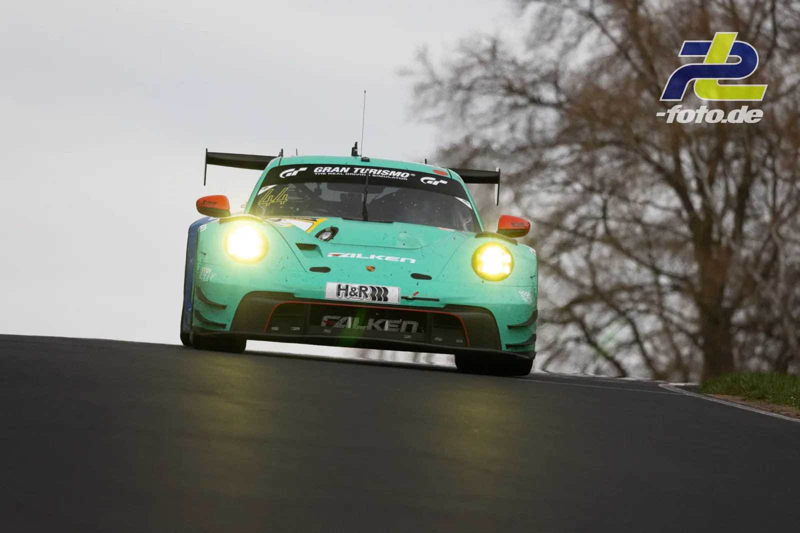 Porsche 911 GT3 R in türkiser Falken-Lackierung über Kuppe fliegend auf der Nordschleife, Scheinwerfer eingeschaltet.