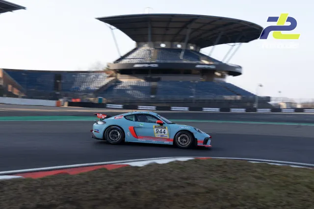 Hellblauer Porsche 718 Cayman GT4 mit Startnummer 944 auf der Nordschleife am Nürburgring, im Hintergrund die Main-Tribüne.