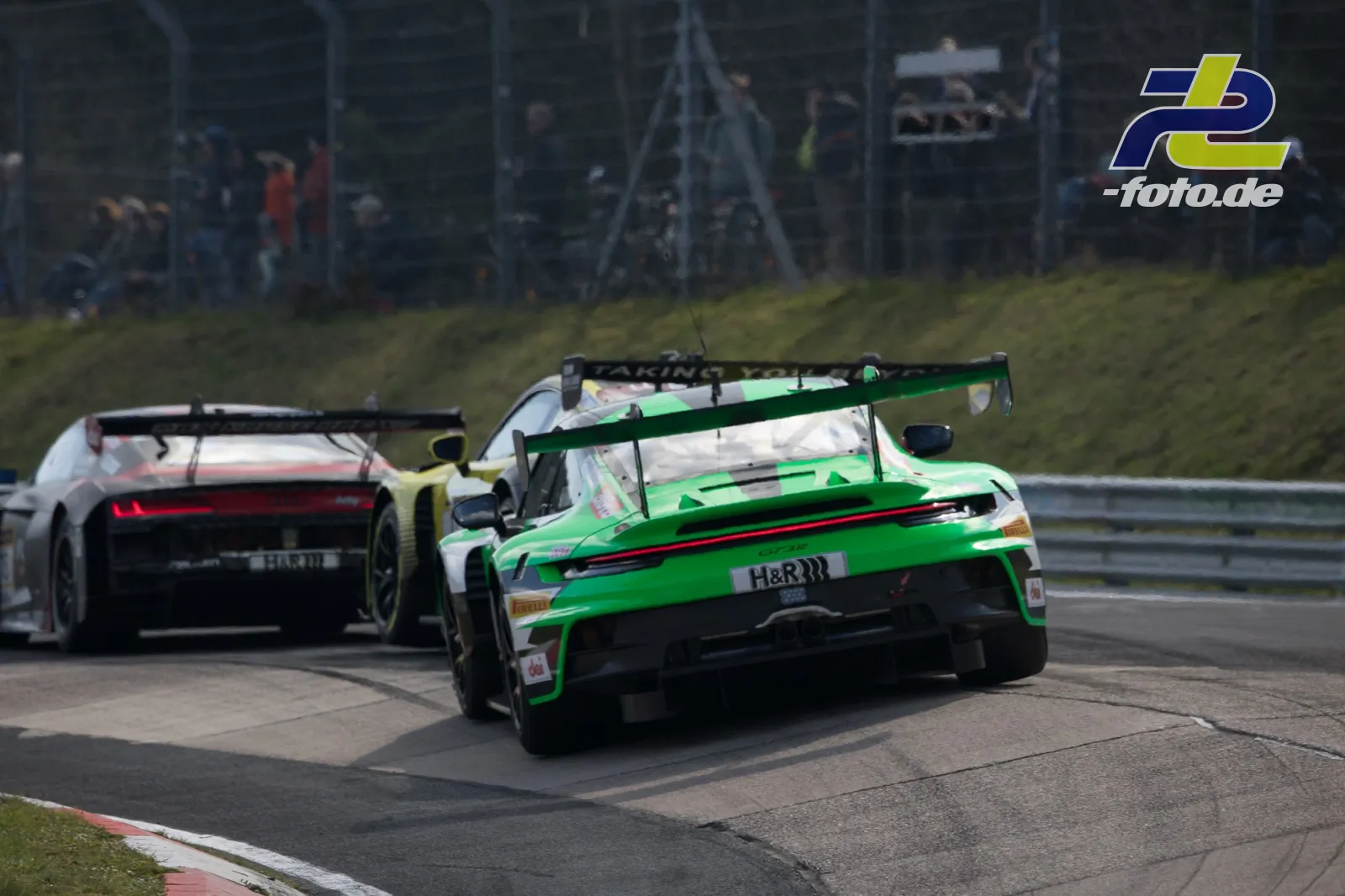 Zwei GT3-Rennwagen in Kurve auf dem Nürburgring, grün-weißer Porsche vor gelbem Fahrzeug mit Zuschauern im Hintergrund.