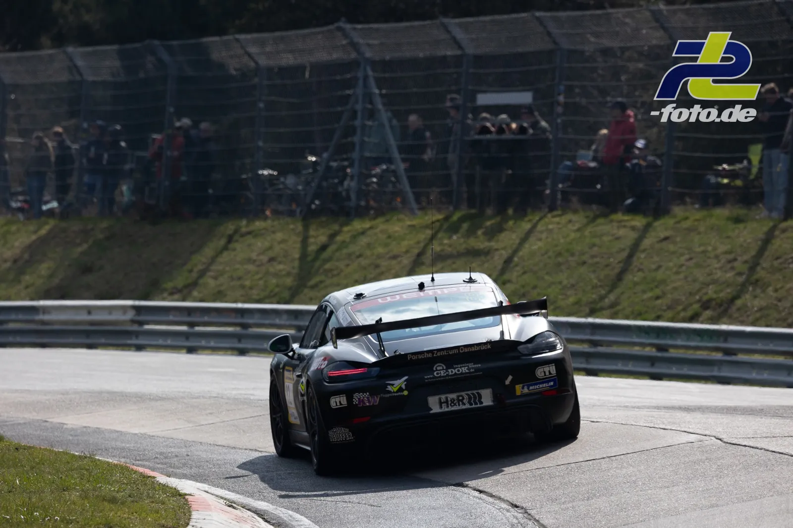 Porsche 718 Cayman GT4 in schwarz-weißer Lackierung während des Rennens am Nürburgring, Heckansicht mit Zuschauern im Hintergrund