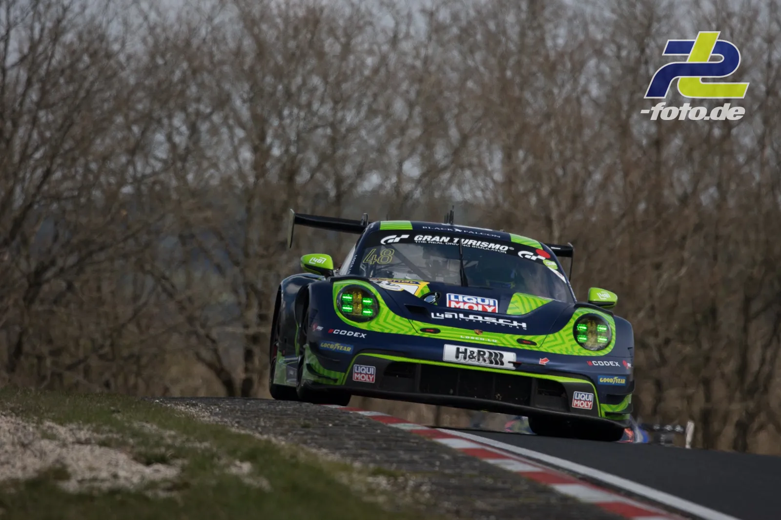 Porsche 911 GT3 R in neongrün-schwarzer Lackierung springt über die Kuppe der Nordschleife, kahle Bäume im Hintergrund.