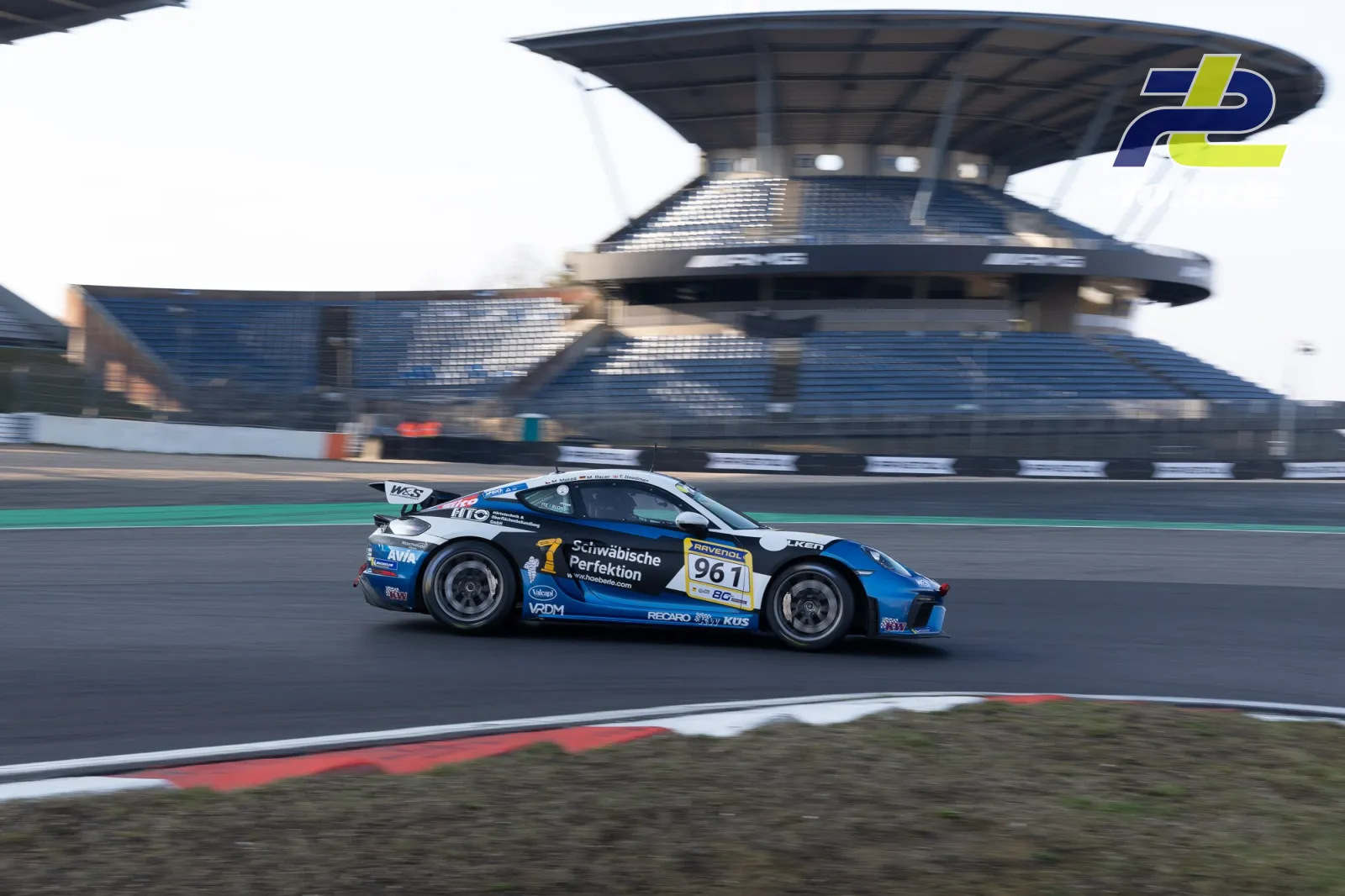 Porsche Cayman GT4 #961 in blau-schwarzer Lackierung fährt im Zeittraining auf dem Nürburgring, Tribüne im Hintergrund.