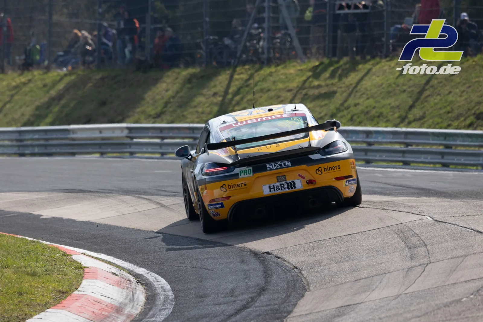 Porsche 718 Cayman in gelb-schwarzer Lackierung in der Kurve während des NLS-Rennens auf dem Nürburgring, Zuschauer im Hintergrund.