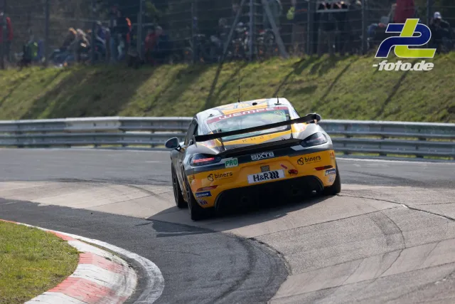 Porsche 718 Cayman in gelb-schwarzer Lackierung in der Kurve während des NLS-Rennens auf dem Nürburgring, Zuschauer im Hintergrund.