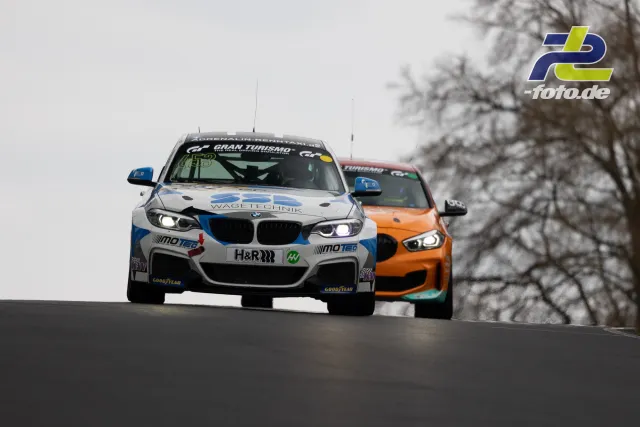 Zwei BMW-Rennwagen im Zweikampf auf der Nürburgring-Nordschleife bei trübem Wetter, im Hintergrund kahle Bäume.