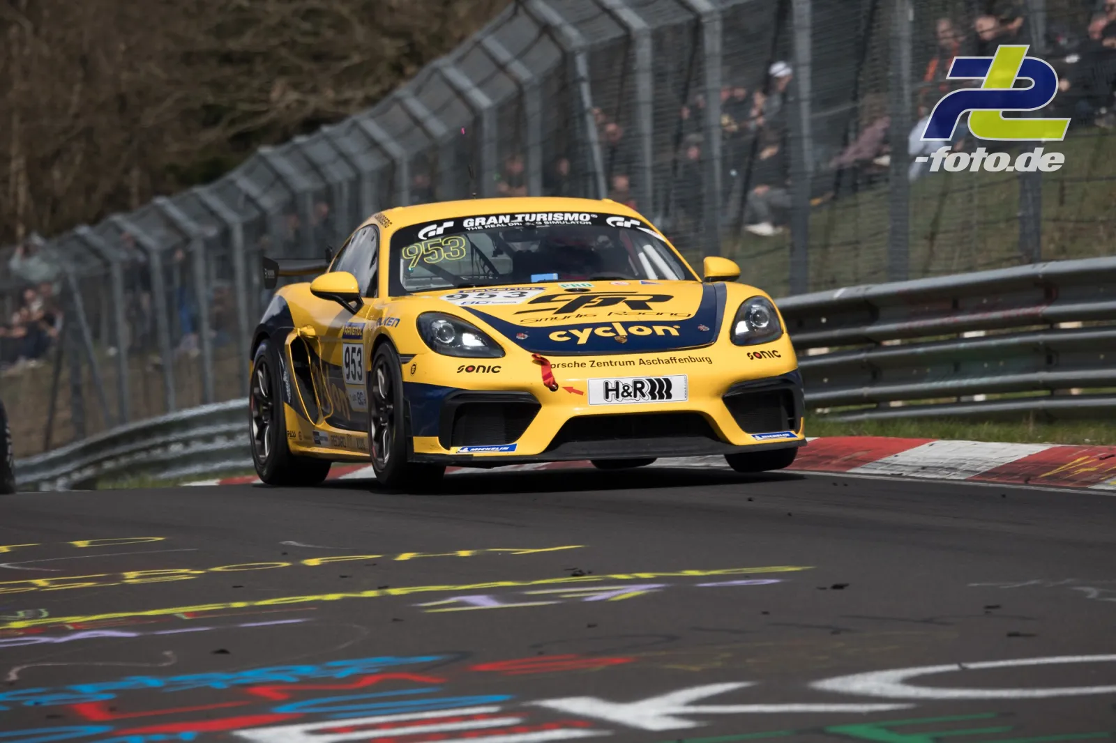 Gelber Porsche 718 Cayman GT4 Clubsport #953 mit cyclon-Lackierung in Kurve auf der Nürburgring-Nordschleife beim NLS-Rennen.