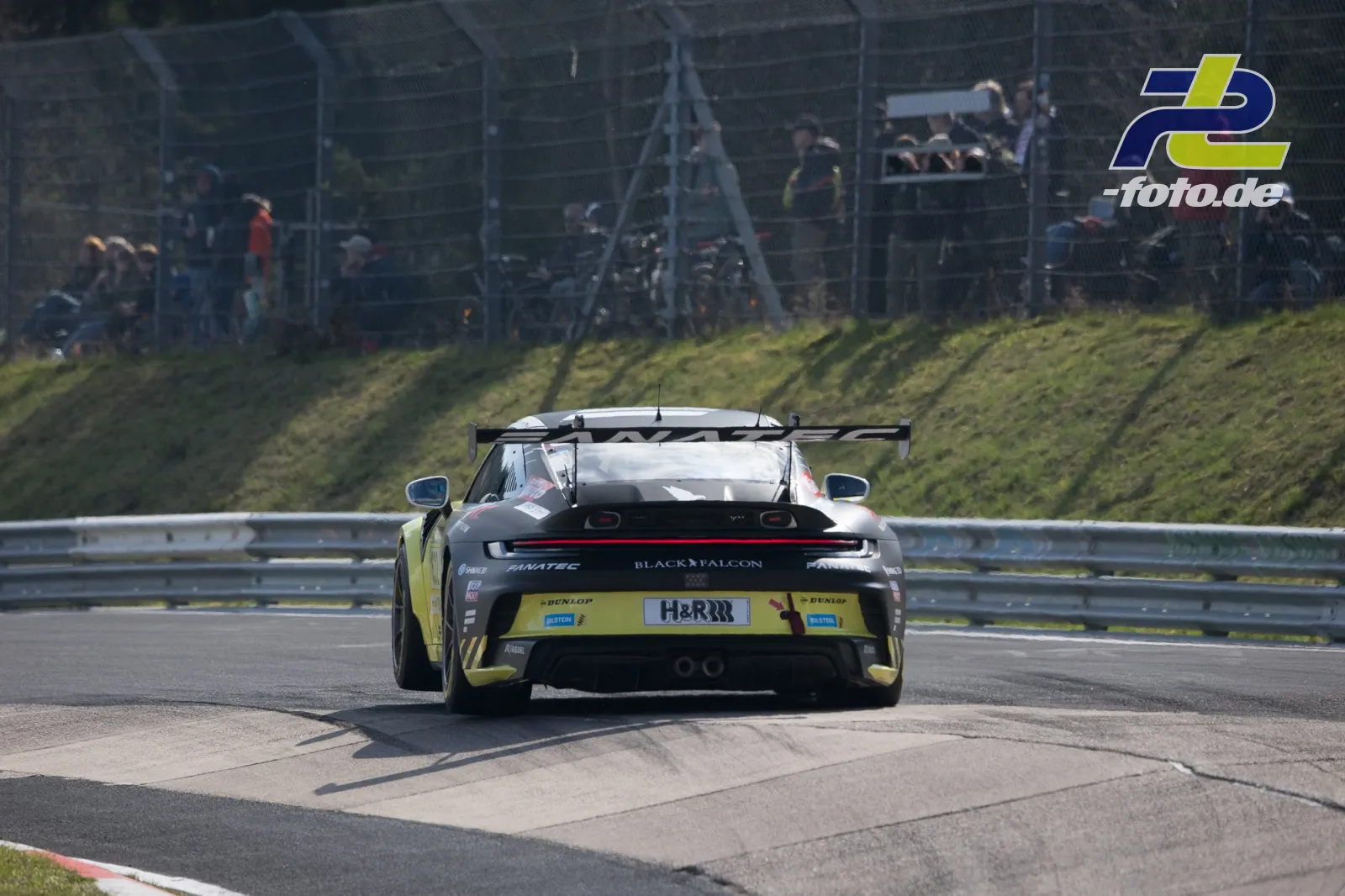 Porsche 911 GT3 in schwarz-gelber Lackierung von hinten auf der Nordschleife, Zuschauer am Streckenrand.