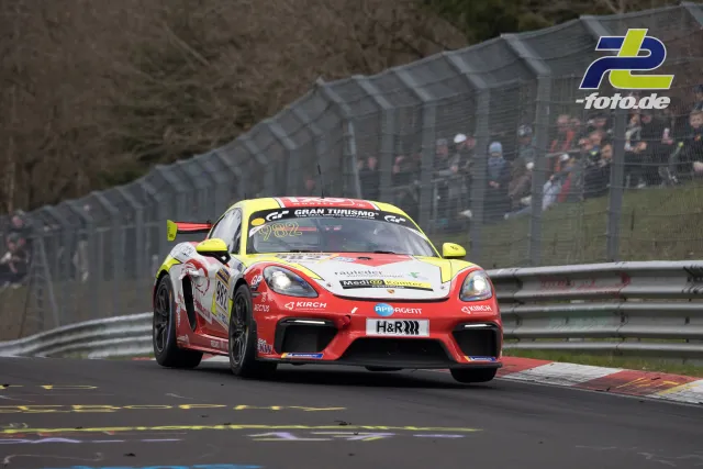 Porsche 718 Cayman GT4 in rot-gelb-weißer Lackierung auf der Nordschleife beim NLS-Rennen, Zuschauer am Zaun.
