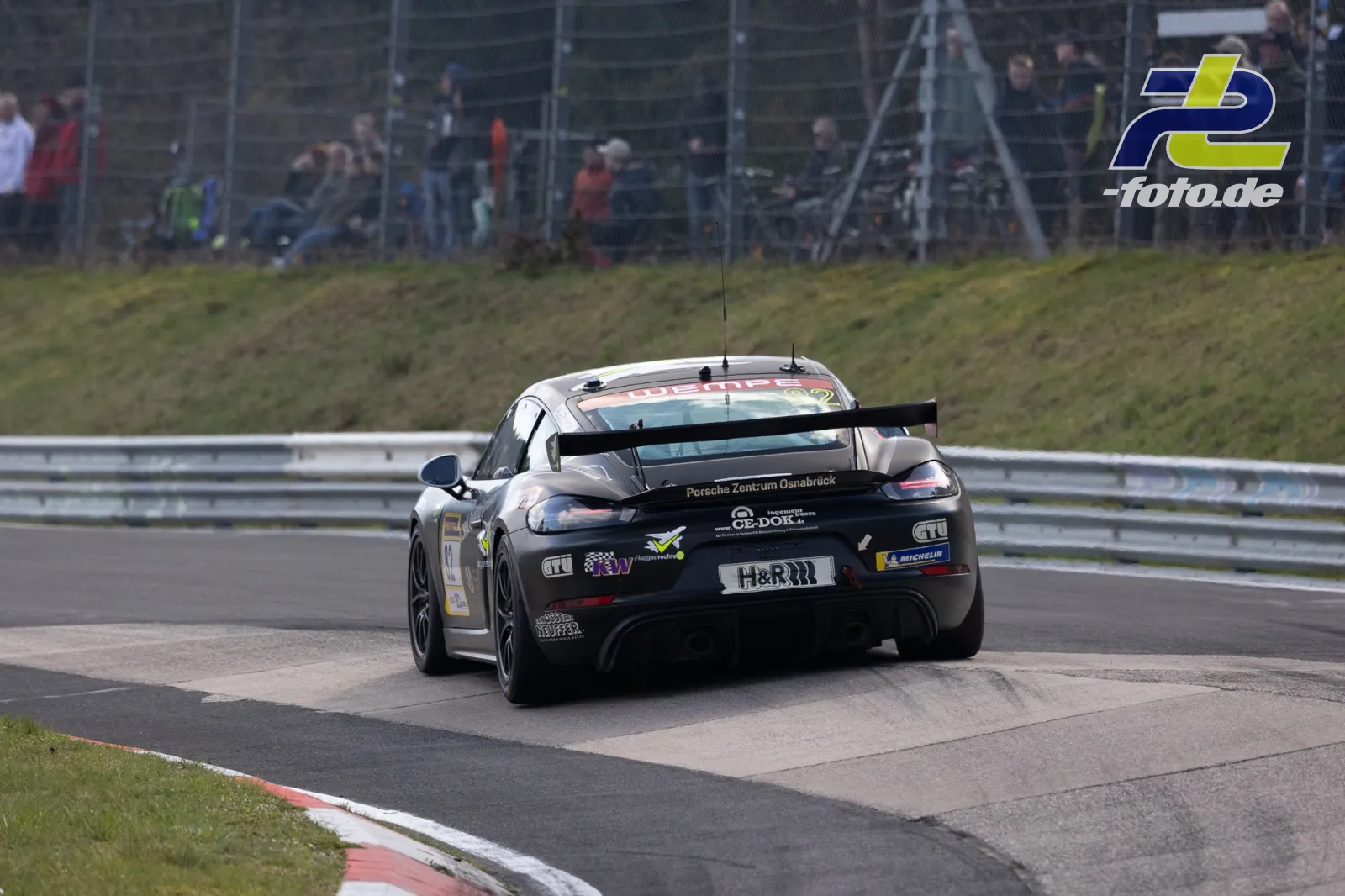 Porsche Cayman GT4 vom Porsche Zentrum Osnabrück in einer Kurve auf dem Nürburgring, Heckansicht mit Zuschauern im Hintergrund.