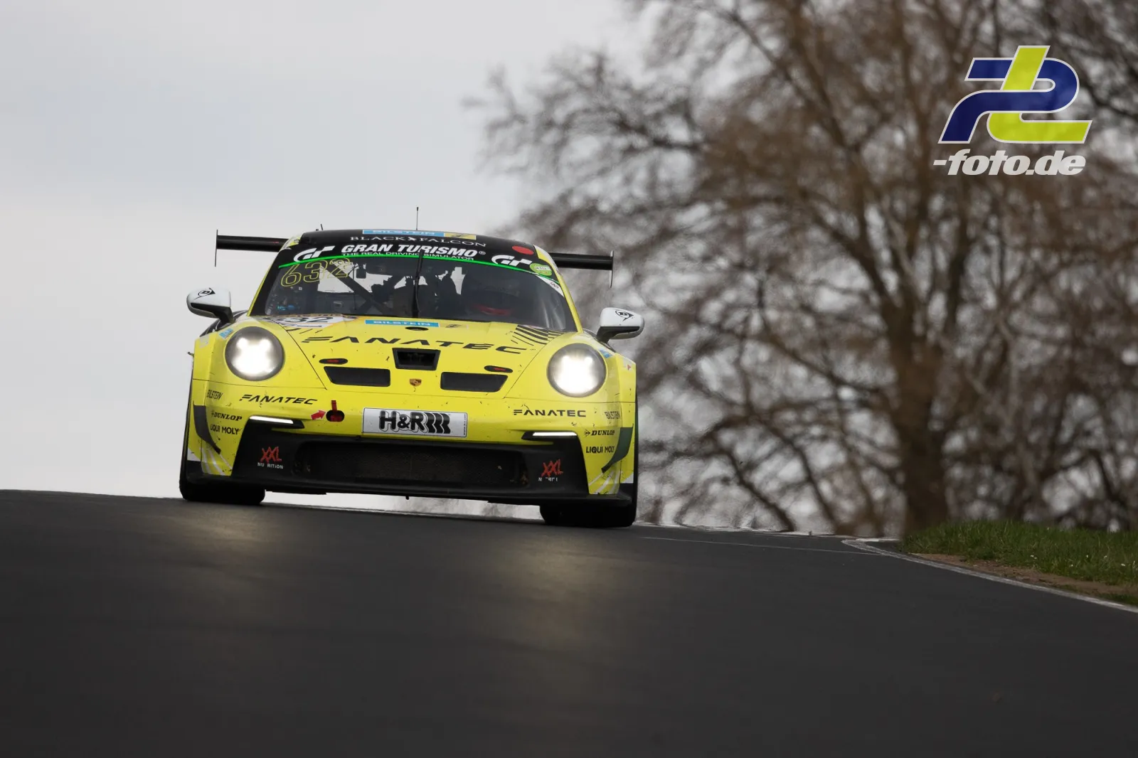 Gelber Porsche 911 GT3 R von Manthey Racing in der Kuppe auf der Nürburgring-Nordschleife bei bewölktem Wetter mit kahlen Bäumen im Hintergrund.